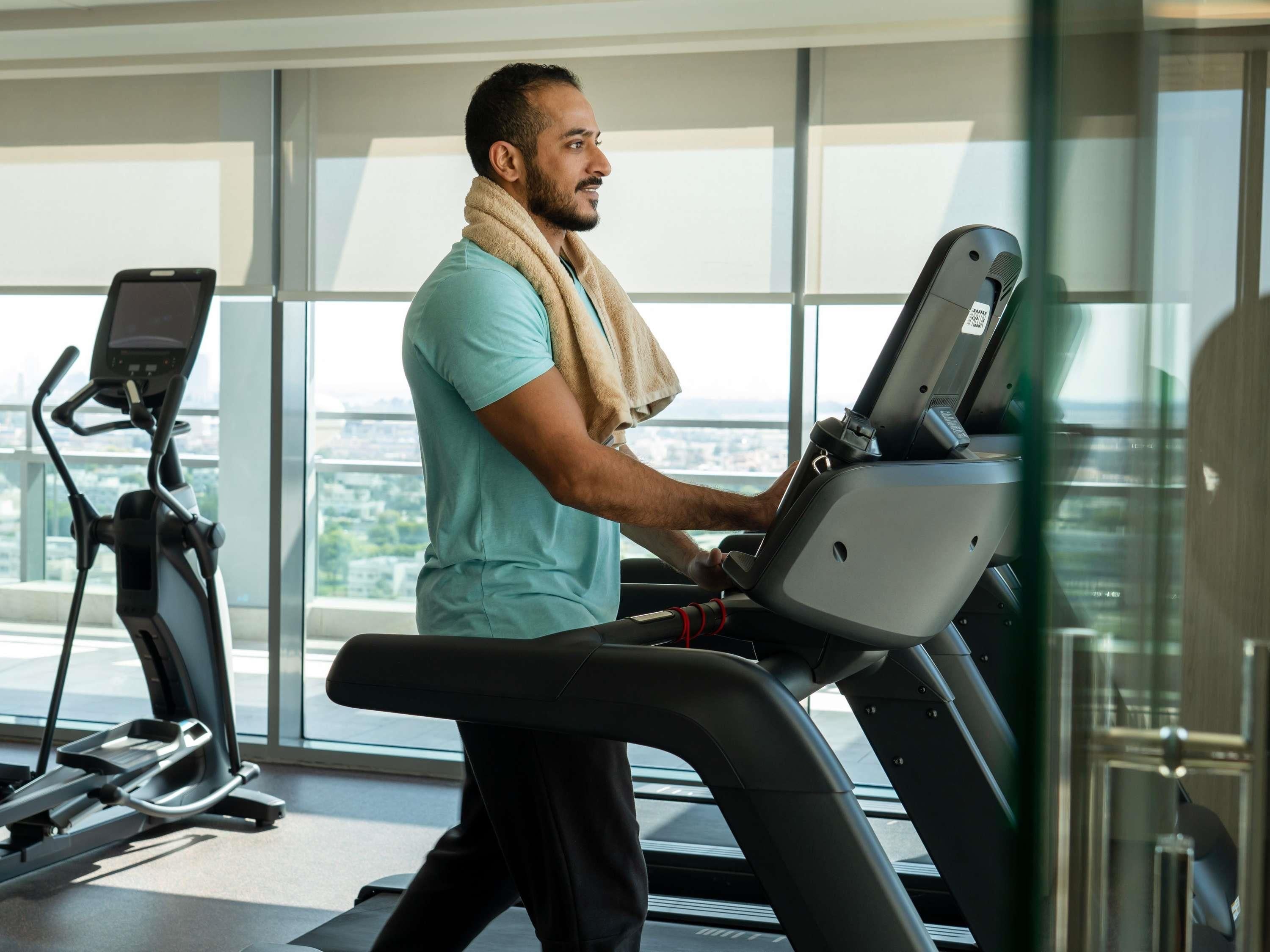 Un hombre usando una caminadora en un gimnasio con ventanas amplias y otra máquina elíptica al fondo.