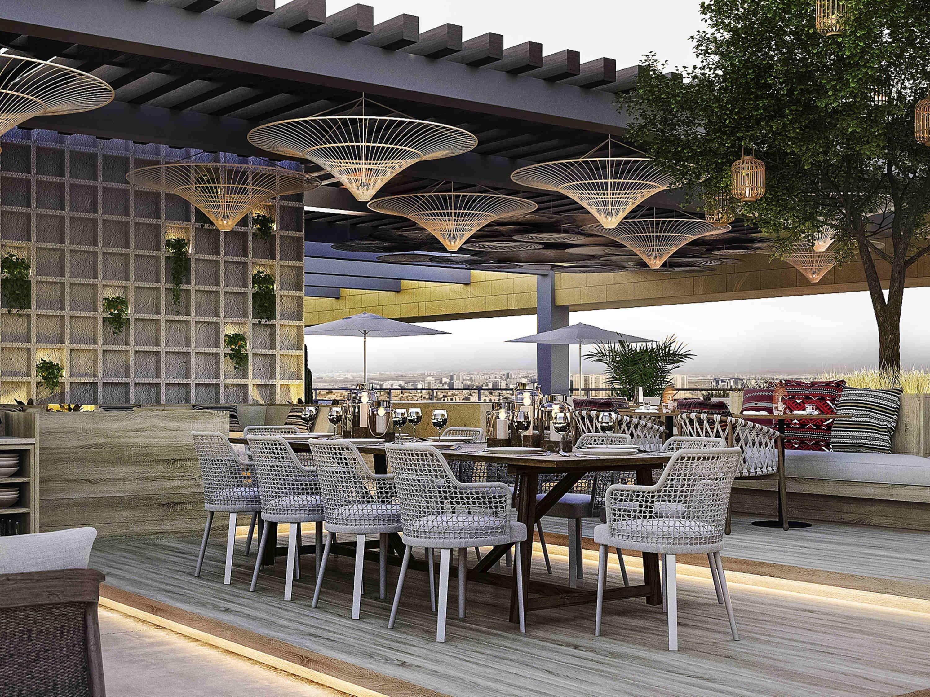 An outdoor dining area with wicker chairs and tables, overhead lighting, and a city view.