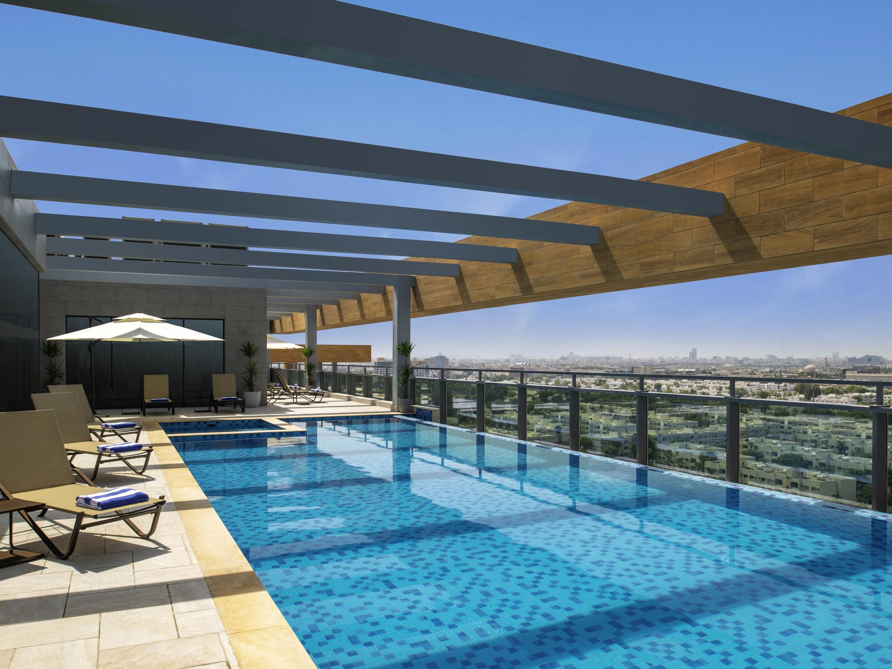 Una piscina infinita en la azotea con sillones de descanso y sombrillas, rodeada por una pérgola de madera y vistas a la ciudad.