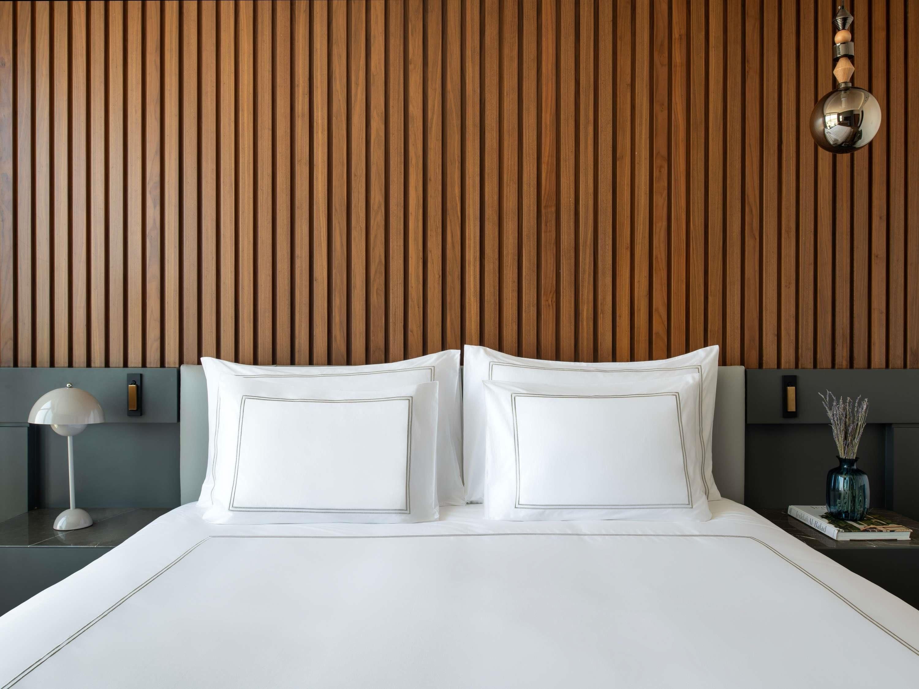 A modern bedroom with a wooden slat headboard, a white bed with two pillows, a bedside table with a lamp, and a small vase with dried flowers.