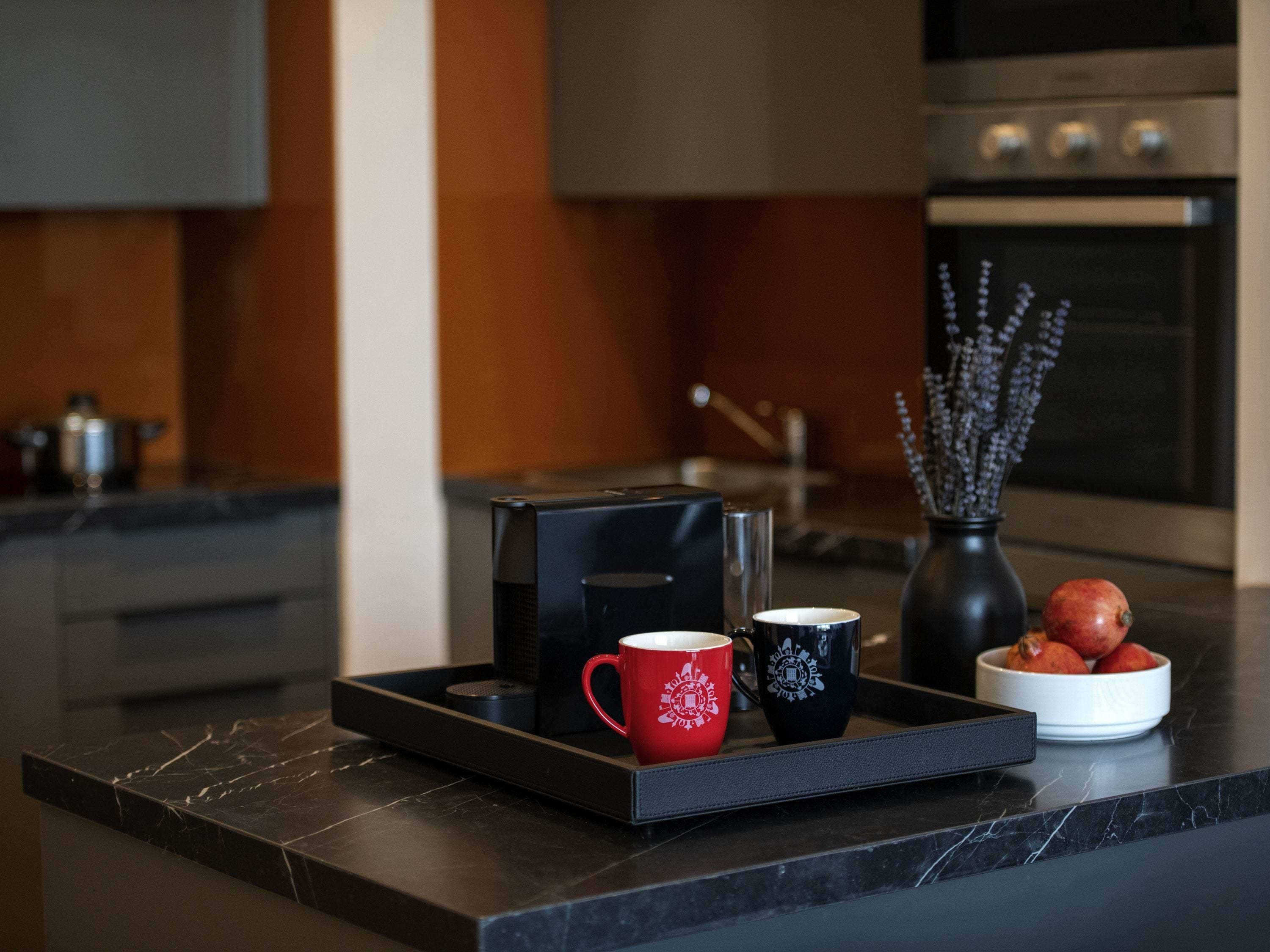 Una cocina moderna con encimera negra, una cafetera, dos tazas, un florero con flores y un cuenco de fruta.