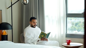 A person reading a book in a well-lit room with a bed, a small table with a cup, and a view of greenery outside. - Swissotel Living Jeddah (Jeddah)