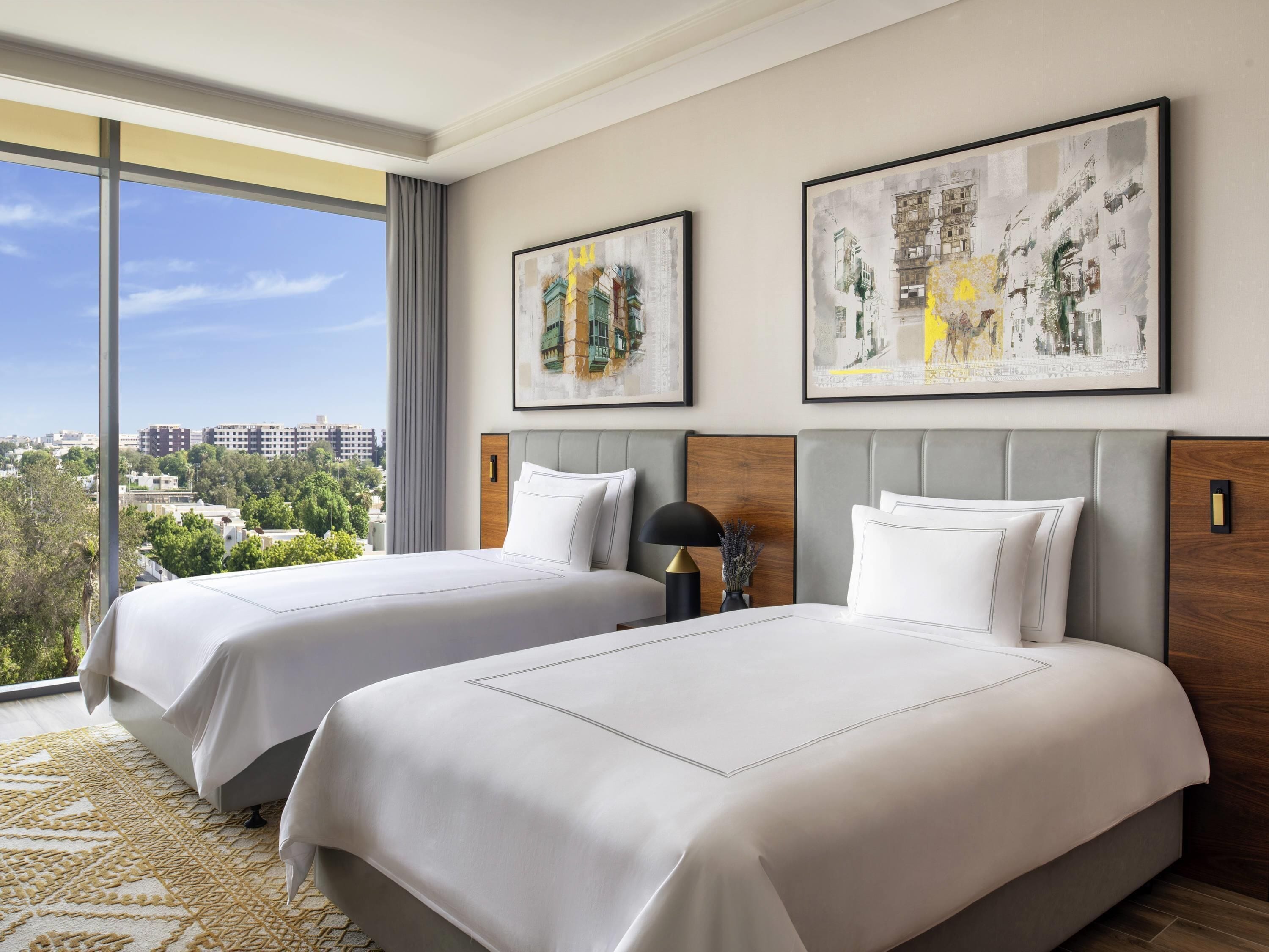 Une chambre d’hôtel avec deux lits, une grande fenêtre donnant sur la ville et deux œuvres d’art encadrées accrochées au mur.