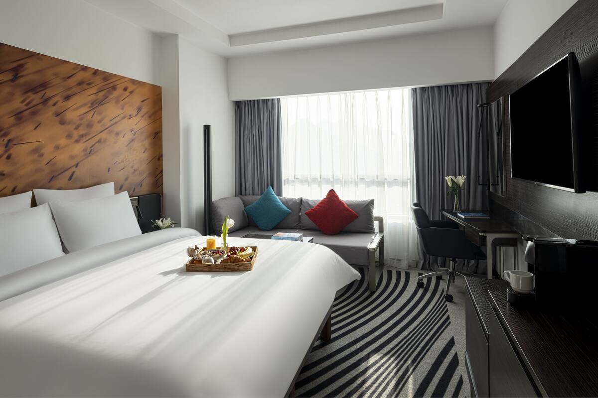 A modern hotel room with a large bed, a desk, a flat-screen TV, and a large window with curtains.