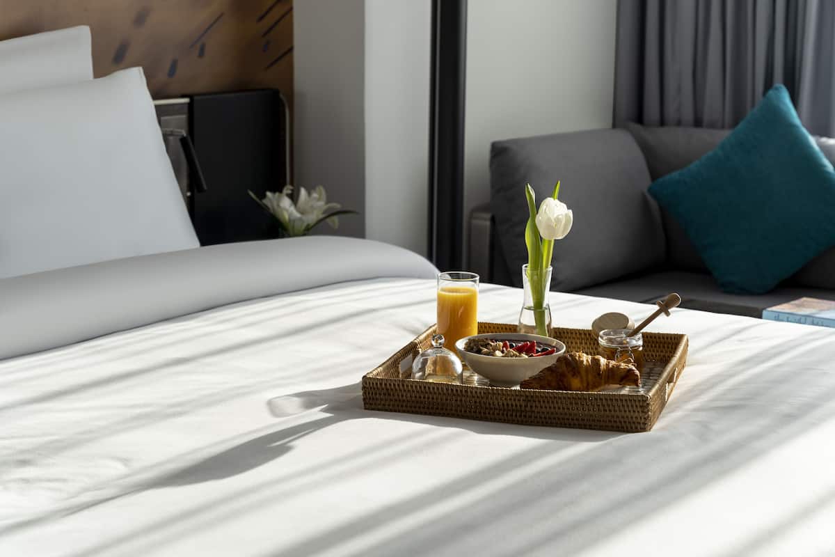 A neatly made breakfast tray on a bed with a croissant, bowl of cereal, and glass of orange juice.