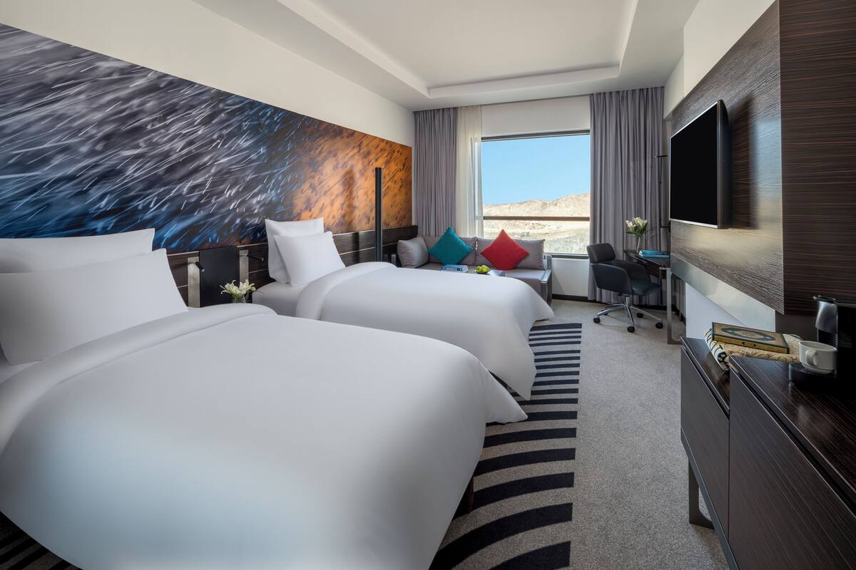 A hotel room with two beds, a desk, a chair, a TV, and a large window with a desert view.