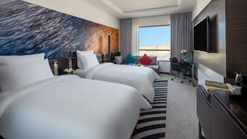 A hotel room with two beds, a desk, a chair, a TV, and a large window with a desert view.