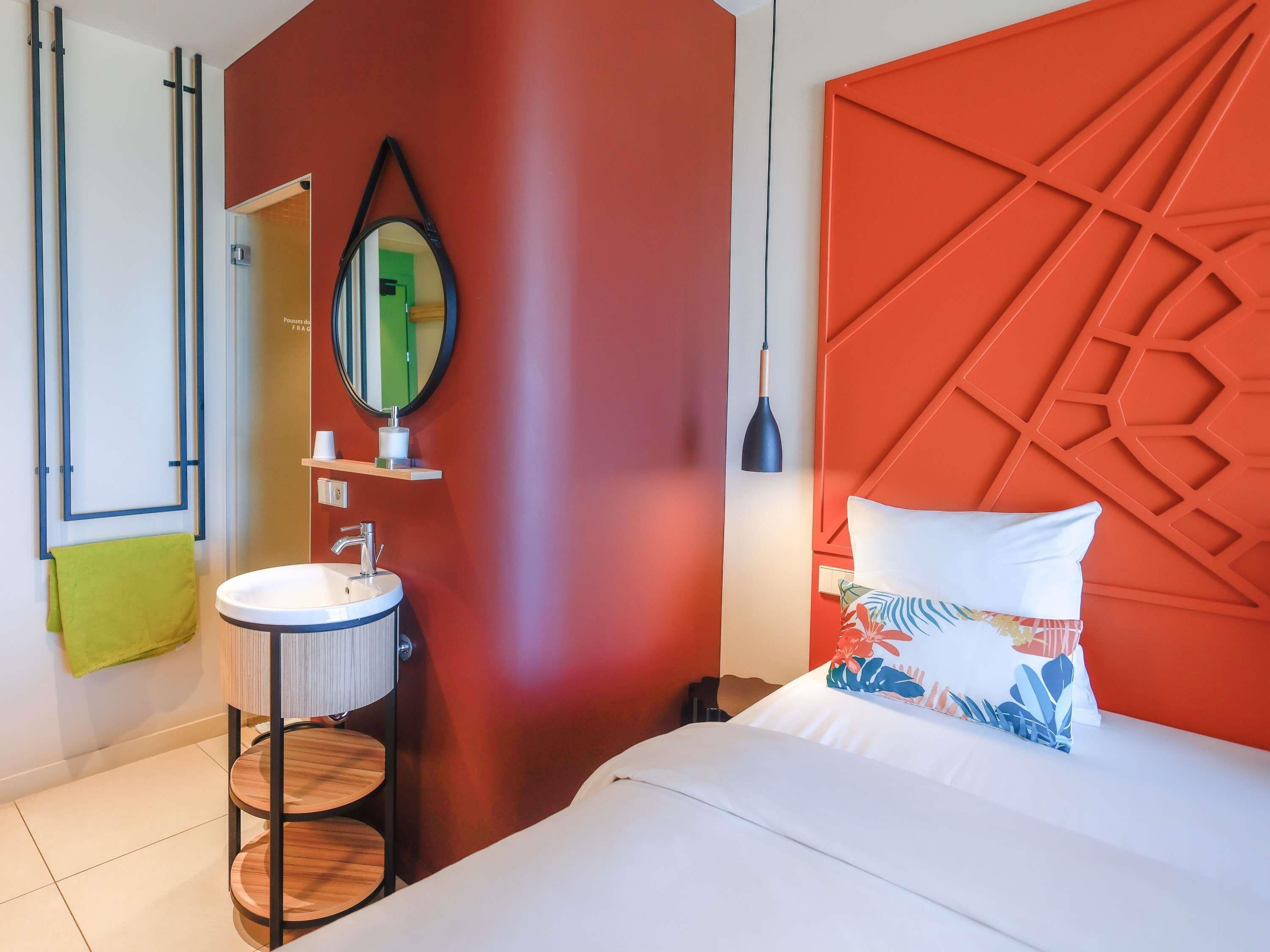 A modern hotel room with a round sink, a mirror, a bed with a geometric headboard, and a green towel.