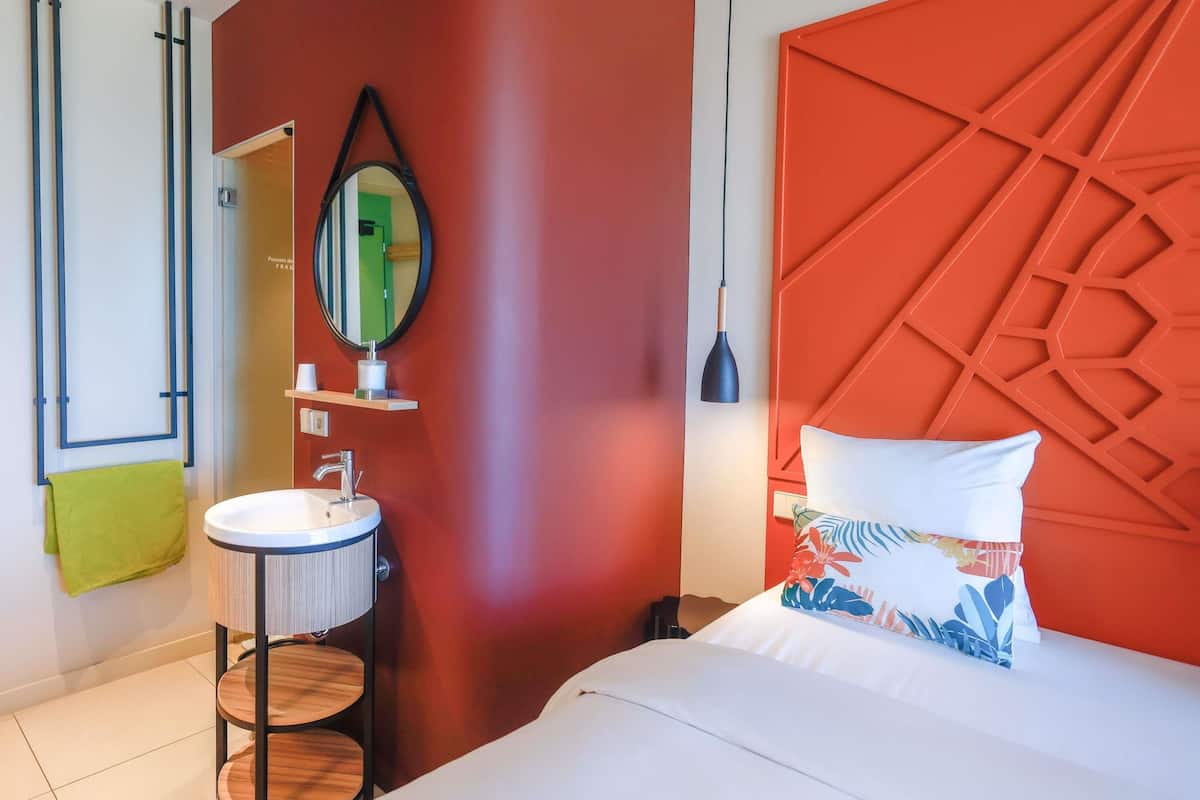 A modern hotel room with a round sink, a mirror, a bed with a geometric headboard, and a green towel.