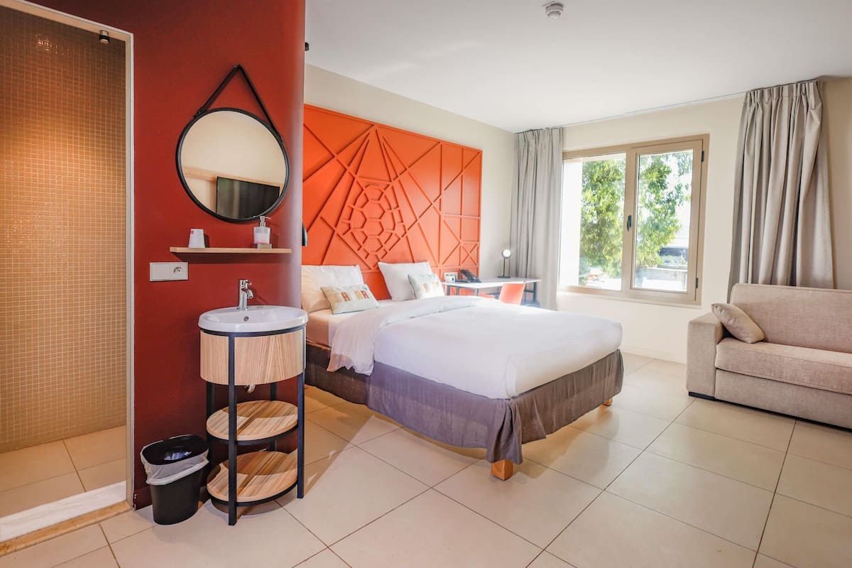 A hotel room with a bed, a round mirror, a sink, a chair, and a window with curtains.
