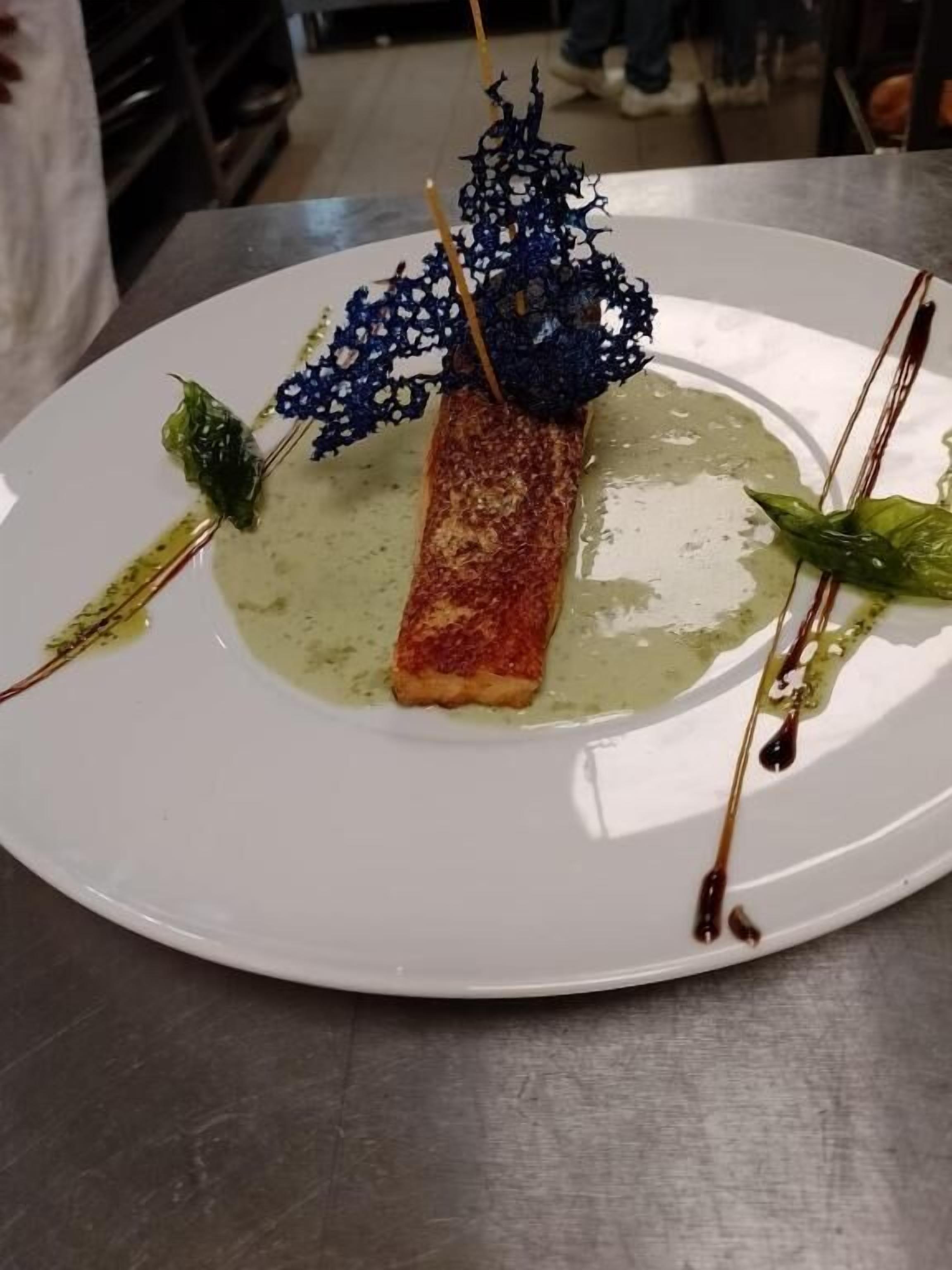 A plate with a piece of food, possibly salmon, topped with a decorative blue structure and garnished with green leaves.