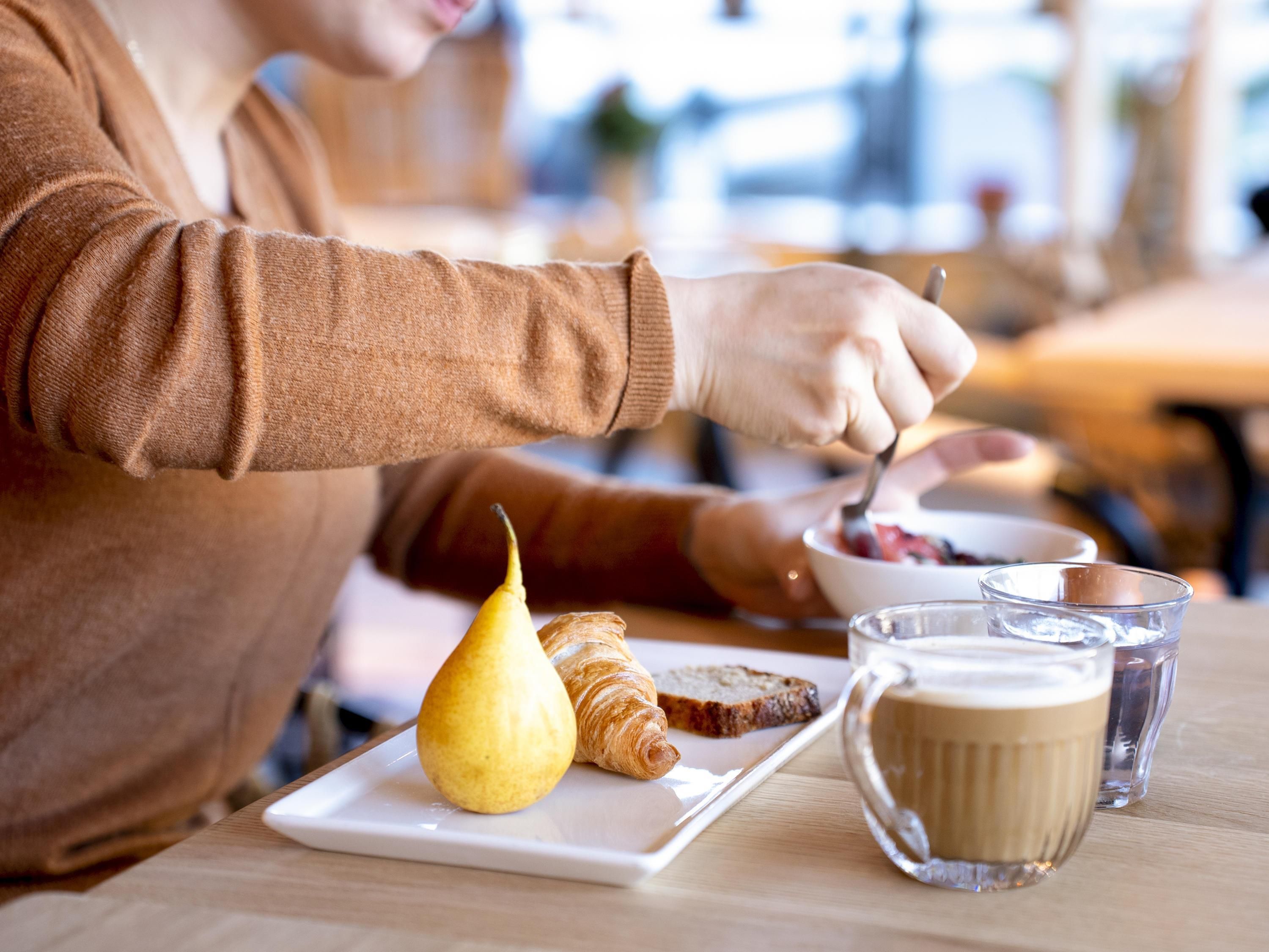 A person eating breakfast with croissants, a pear, and coffee on a wooden table.