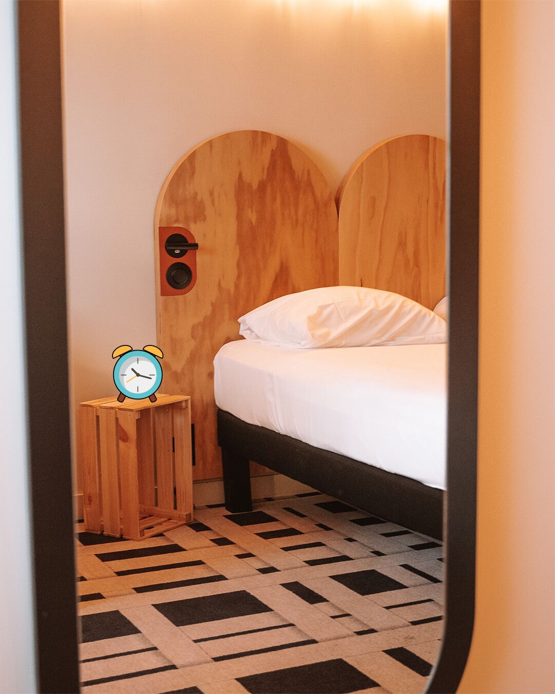 A small, cozy room with a bed, a wooden headboard, a nightstand with a clock, and a patterned floor.