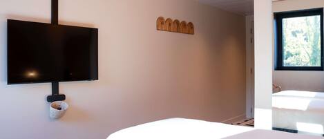 A hotel room with a flat-screen TV mounted on the wall, a wooden headboard, a window with a view of trees, and a white bed.