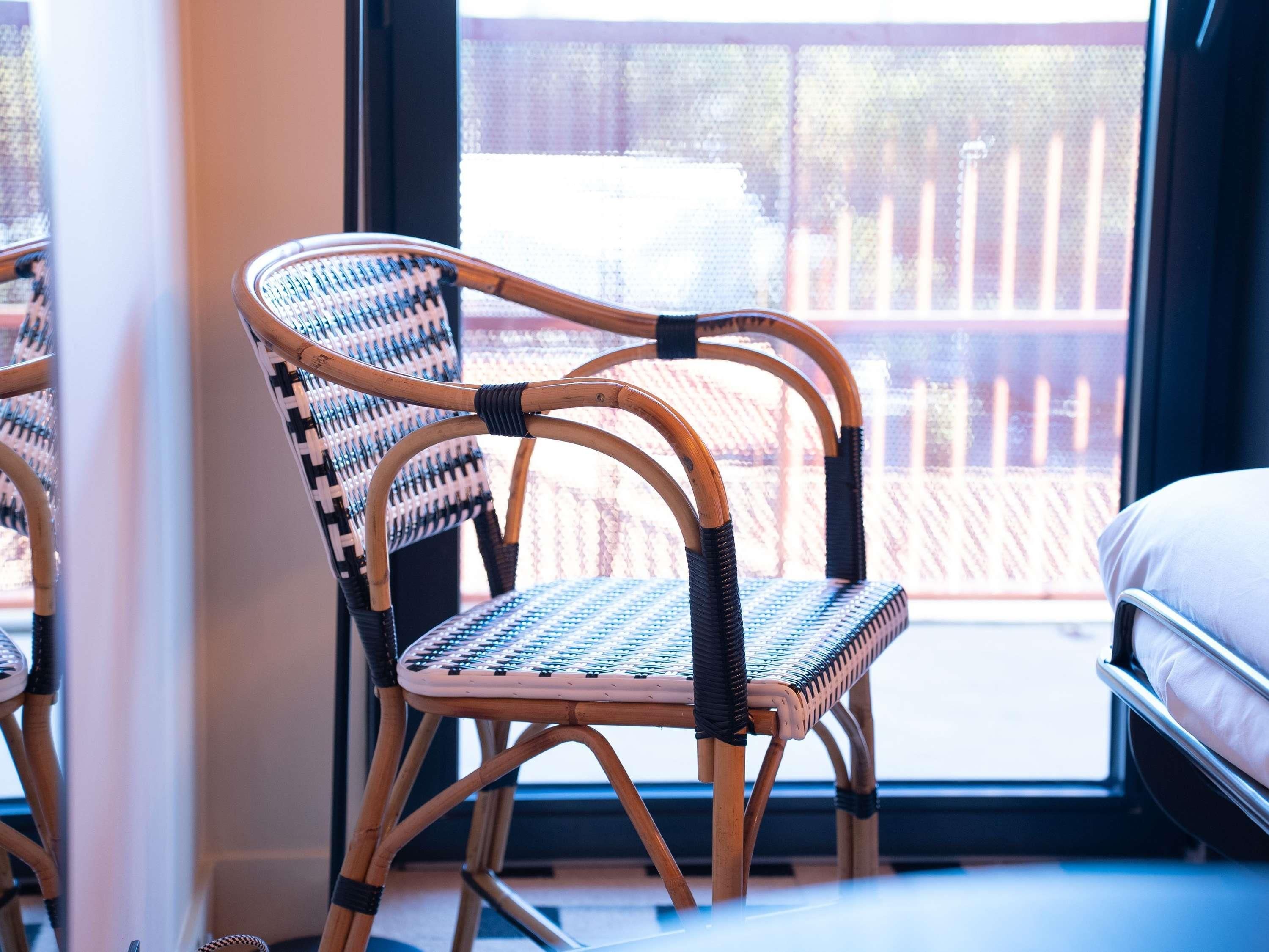 A wicker chair with a woven seat and backrest, positioned near a window with a view of a fence outside.