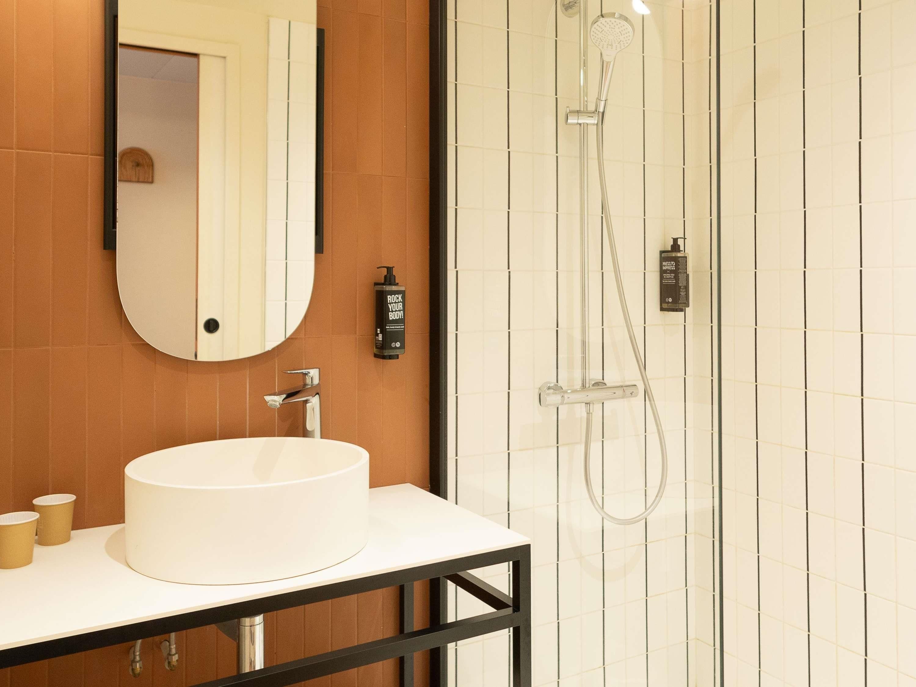 Une salle de bain avec un lavabo blanc, un miroir rond, une douchette à main et une bouteille de shampoing.