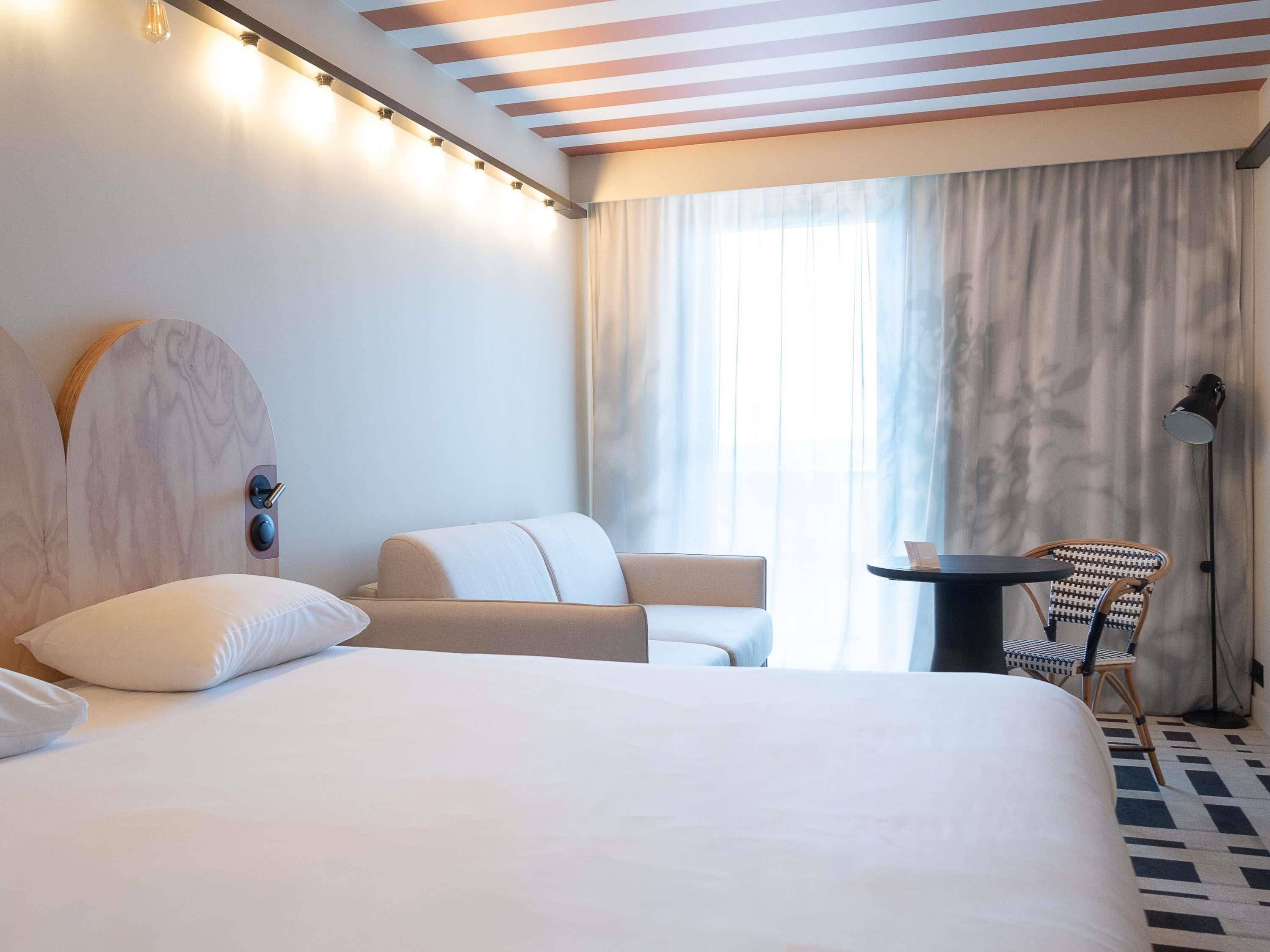Una habitación de hotel con una cama, un escritorio, una silla y un amplio ventanal con cortinas.