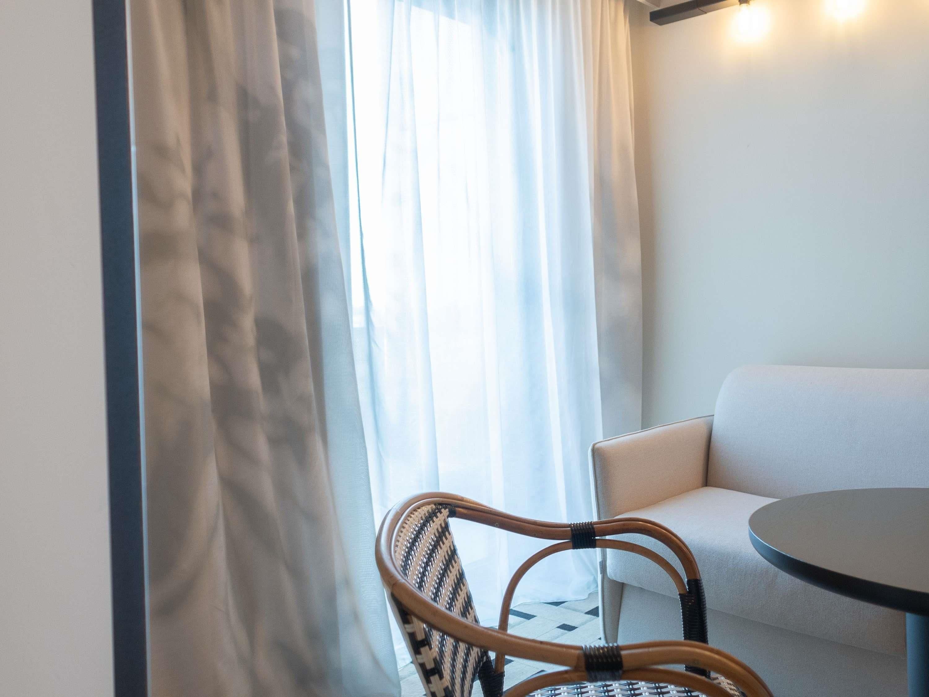 Ein Zimmer mit einem runden Tisch, einem Korbstuhl, einer Couch und einem Fenster mit durchsichtigen Vorhängen.