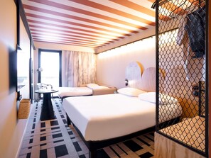 Quarto de hotel com cama, uma mesinha, uma cadeira, janela com cortinas e vista para o exterior.