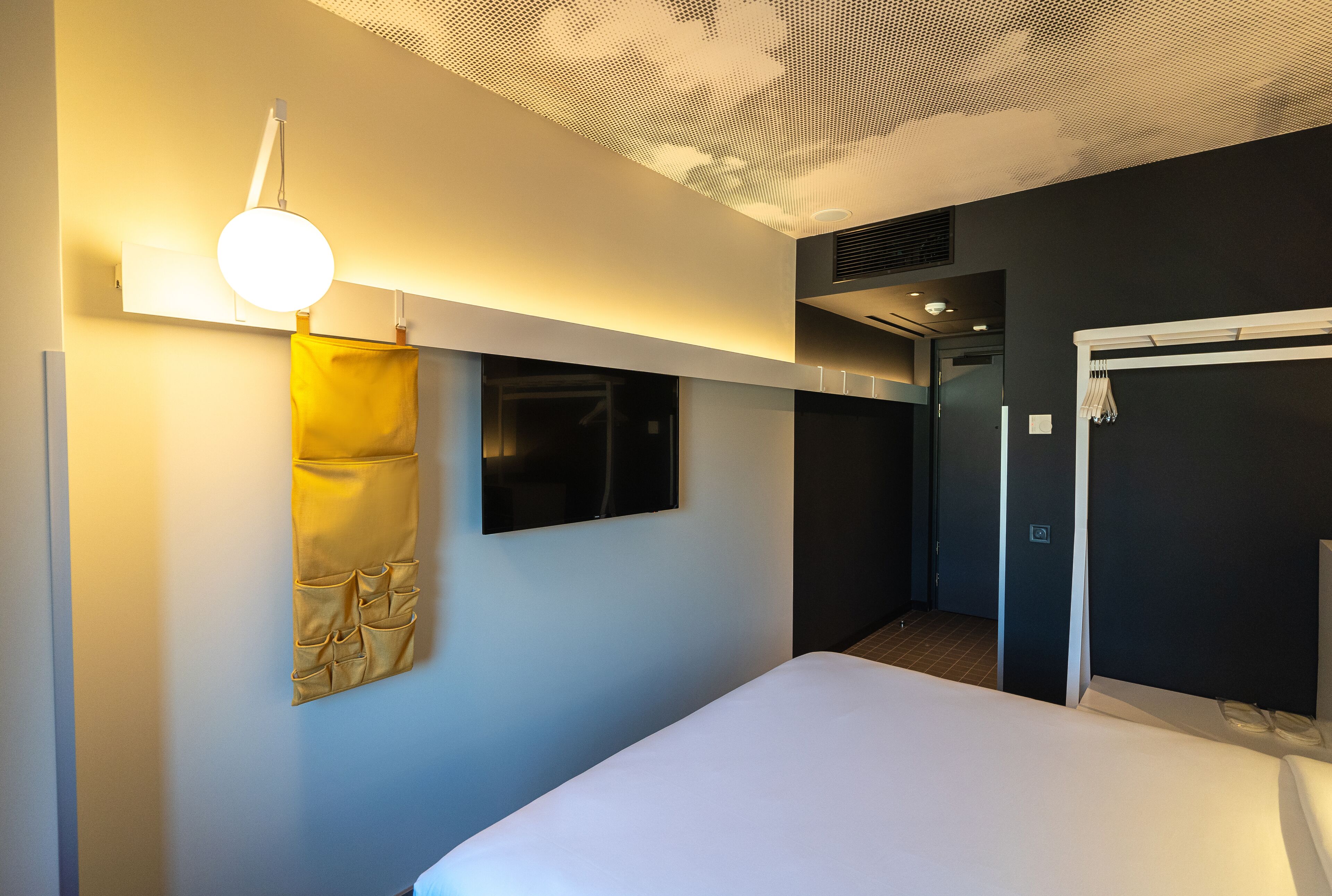 A modern hotel room with a bed, a wall-mounted TV, a hanging yellow towel, and a ceiling with a perforated design.