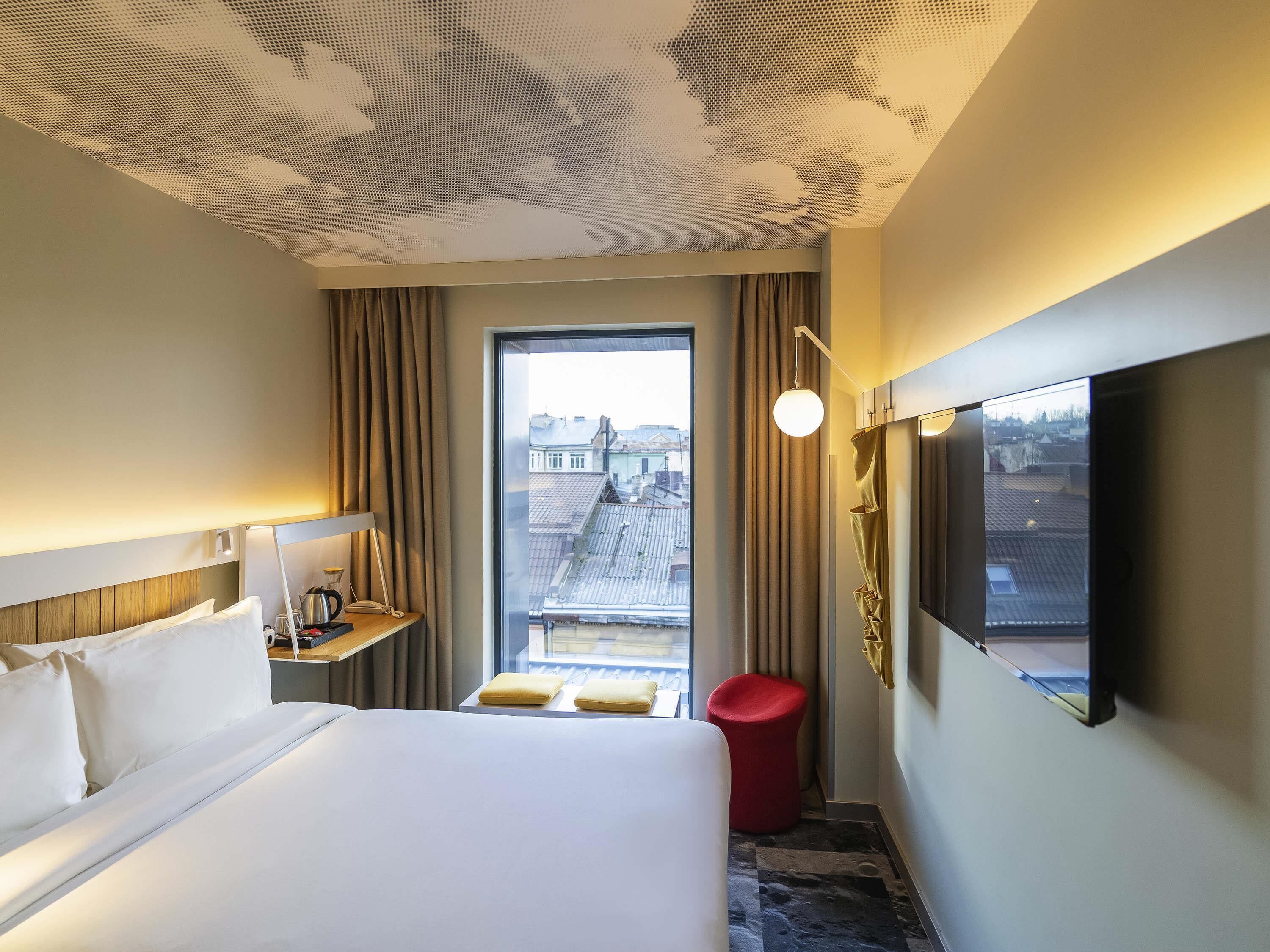 A hotel room with a large bed, a desk with a lamp, a TV mounted on the wall, and a window with a view of buildings.