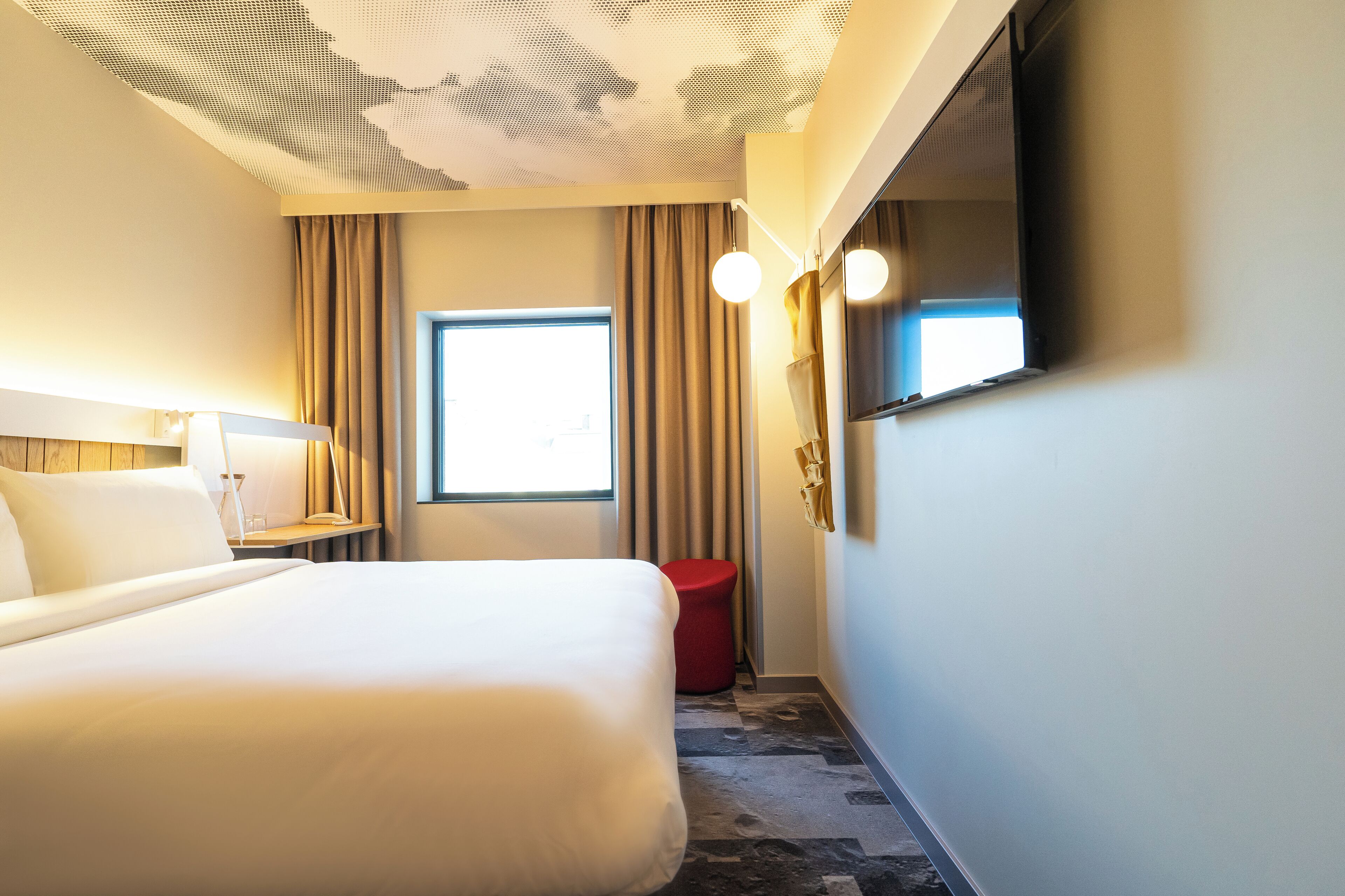A hotel room with a large bed, a TV mounted on the wall, a small table with a lamp, a window with curtains, and a red stool.