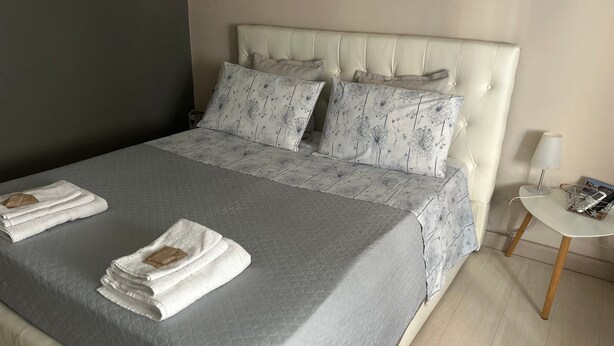 A neatly made bed with a grey quilt and white pillows, a small bedside table with a lamp, and a wall-mounted shelf. - Holiday Apartment "Locazione Panta Rei" with A/C & Wi-Fi (Verona)