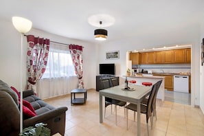Una sala de estar con sofá, una mesa de comedor con sillas, televisor y una cocina con armarios de madera y lavavajillas.