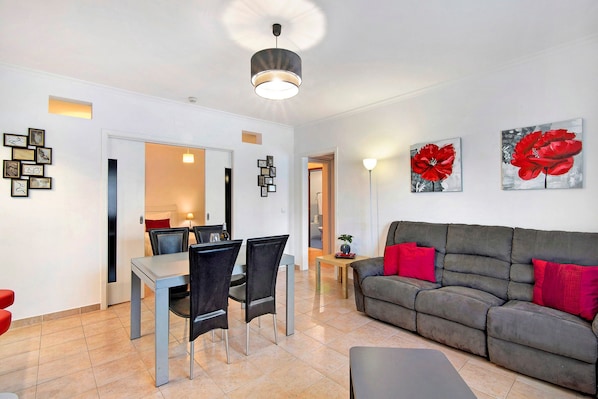 A modern living room with a grey sofa, red cushions, a dining table with chairs, and artwork on the walls.