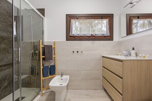 A bathroom with a glass shower enclosure, a bidet, wooden cabinets, a window with a sheer curtain, and a mirror above the sink.