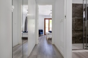 A modern hallway with a glass door leading to a shower area, a blue chair, and a bed with a white headboard.