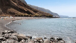 Une plage avec un rivage rocheux, une jetée s’avançant dans des eaux calmes et, en arrière-plan, un paysage montagneux.