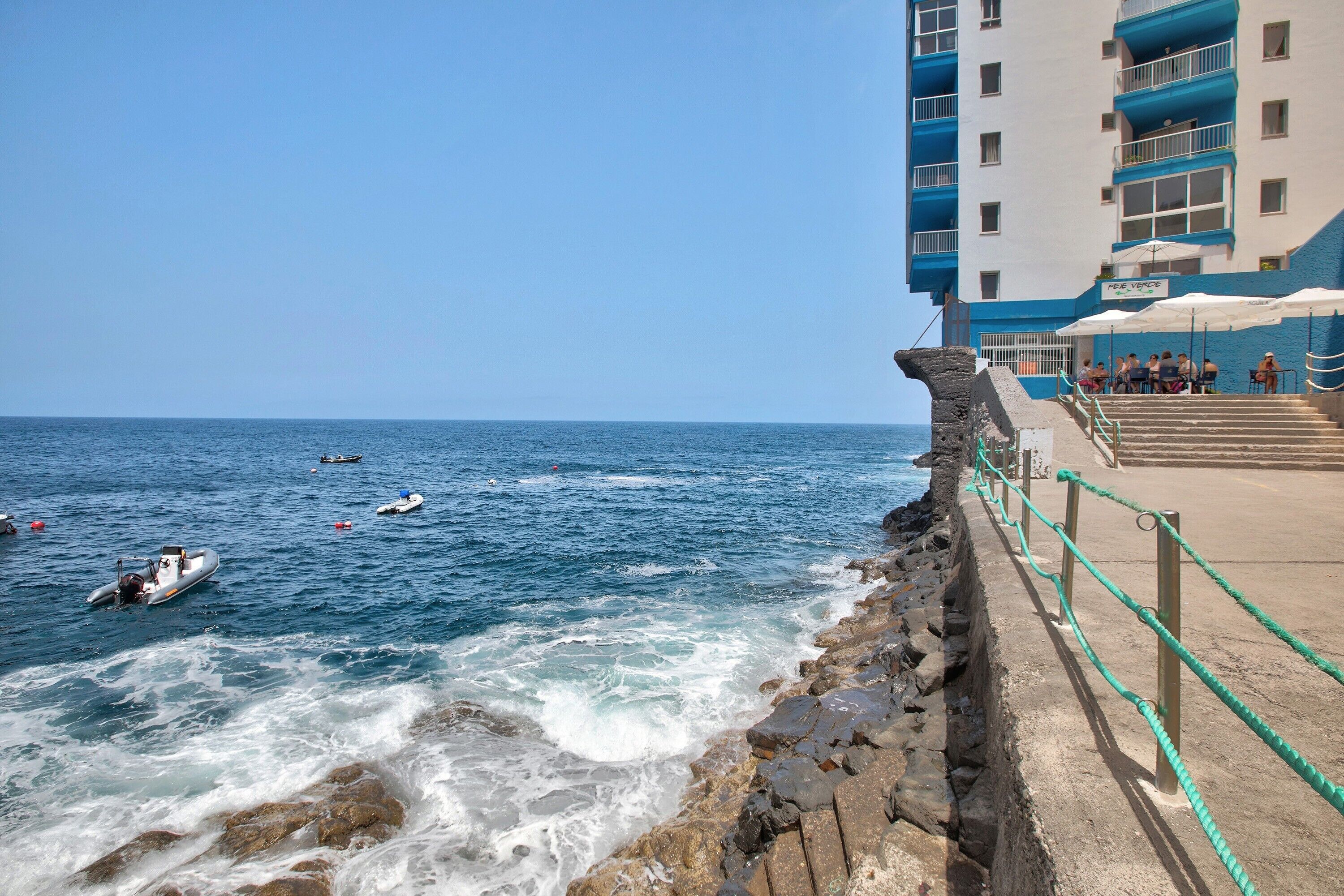 Un hotel costero con un muro de contención de piedra, una zona de piscina con sombrillas y vistas al mar.