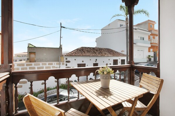 A balcony with a wooden table and chairs, a potted plant, and a view of buildings and a car.