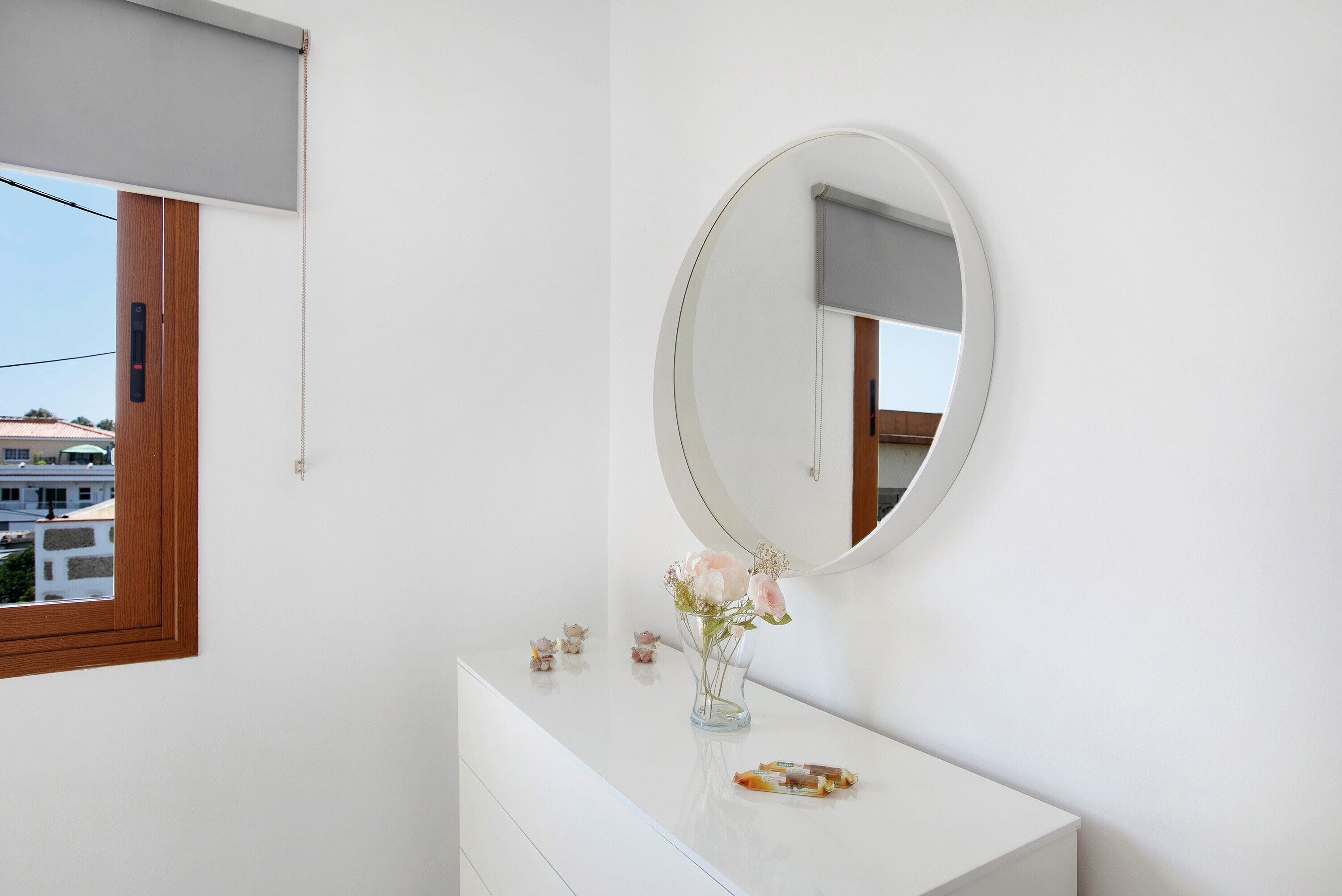 Un baño con tocador blanco, espejo redondo, florero con flores y ventana con vista a edificios.