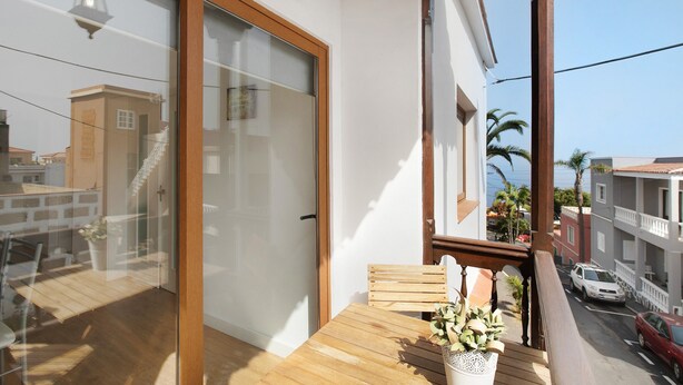 A balcony with a wooden table and chair, a potted plant, and a view of the street and buildings. - Vacation Home 'Casa Micaela 2' with Sea View, Terrace, Balcony & Wi-Fi (El Sauzal)