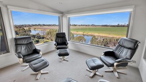 Una habitación con tres sillones de cuero negro y un reposapiés a juego, ventanas amplias con vistas a un lago y campos, y piso alfombrado.