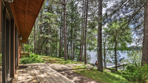 A wooden deck leading to a lakeside cabin with a view of tall pine trees and a calm lake. - Stepaway Vacation Rentals- Schneider Cabin (Sayner)