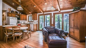 A cabin interior with a wooden ceiling, a dining area with a table and chairs, a leather sofa, and a view of a forest through large windows. - Stepaway Vacation Rentals- Schneider Cabin (Sayner)