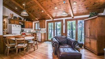 A cabin interior with a wooden ceiling, a dining area with a table and chairs, a leather sofa, and a view of a forest through large windows.
