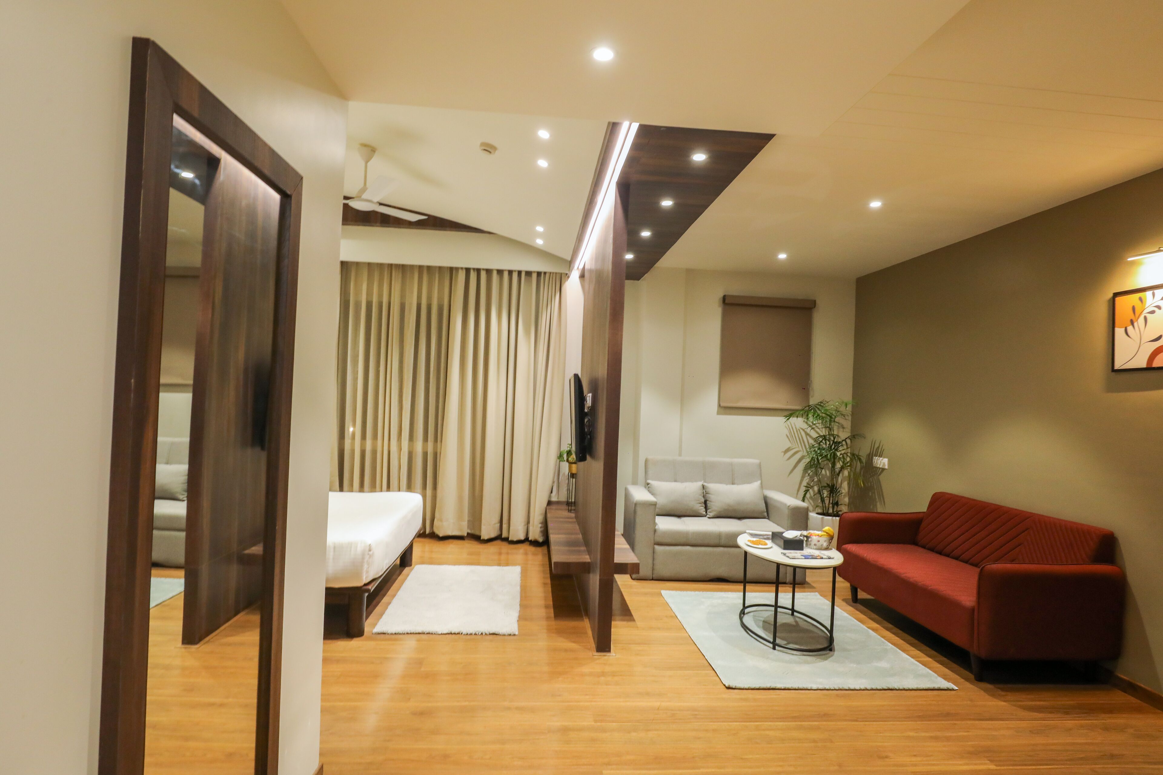 Una habitación de hotel moderna con piso de madera, una cama, un sofá, un sofá rojo, una mesita y una planta en maceta.