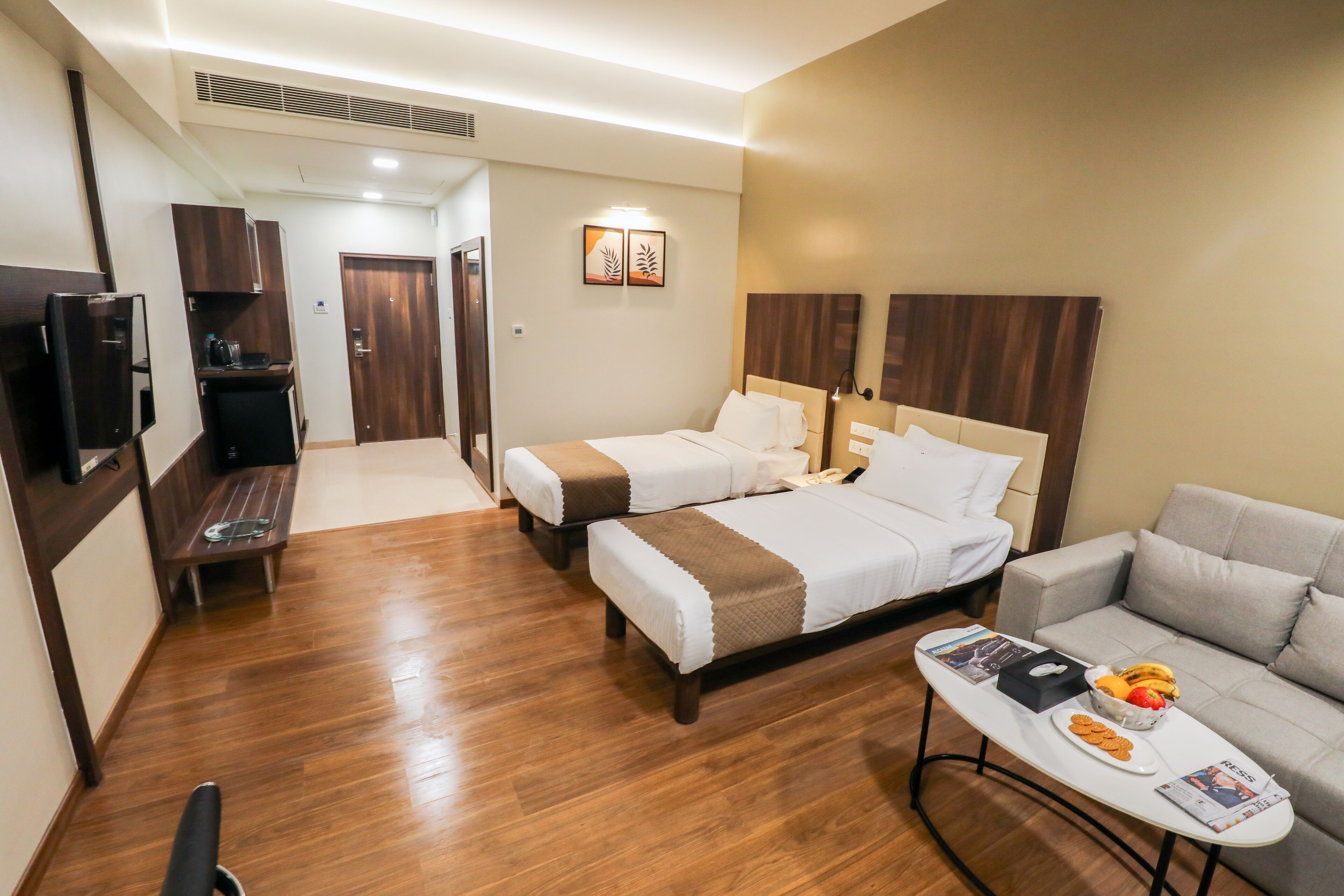 A hotel room with two beds, a sofa, a small table with breakfast items, a TV, and a desk with a computer and coffee machine.