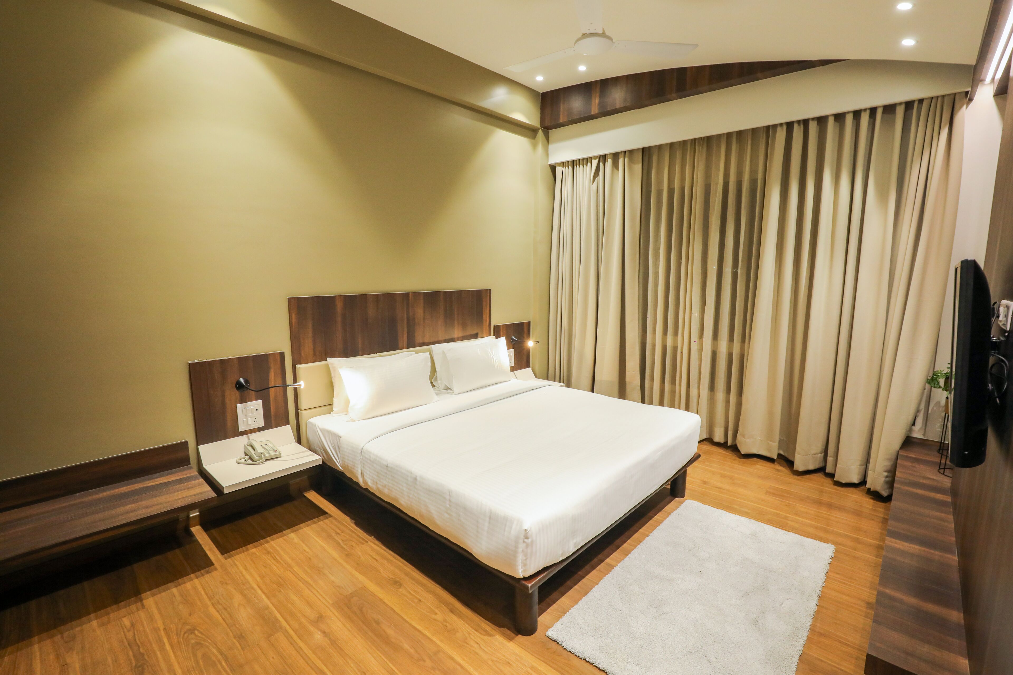 Ein Hotelzimmer mit einem großen Bett, einem Holz-Kopfschild, einem Nachttisch mit Lampe und einem großen Fenster mit Vorhängen.