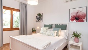 Un dormitorio con cama, una mesita de noche con una planta, una ventana con cortinas y una pared con un cuadro de flores.