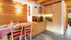 Un intérieur en bois comprenant une table à manger pour deux personnes, un coin cuisine et un fauteuil rouge avec coussin.