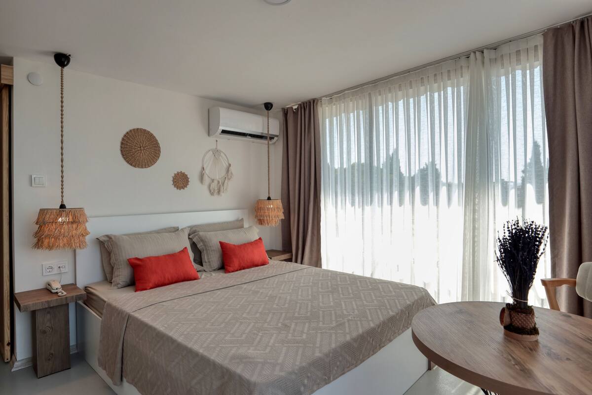A modern bedroom with a large bed, bedside tables, a round wooden table, and a view of the outdoors through sheer curtains.