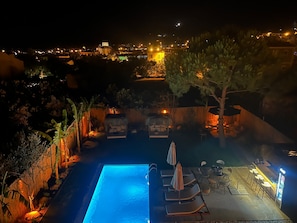 A nighttime view of a swimming pool area with lounge chairs and umbrellas, surrounded by tropical plants and a wooden fence. - Myrodia by Etapaltınel (Ayvalik)