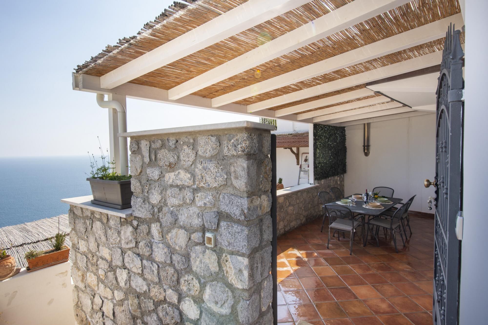 A stone wall with a potted plant, a table with chairs, and a view of the sea.