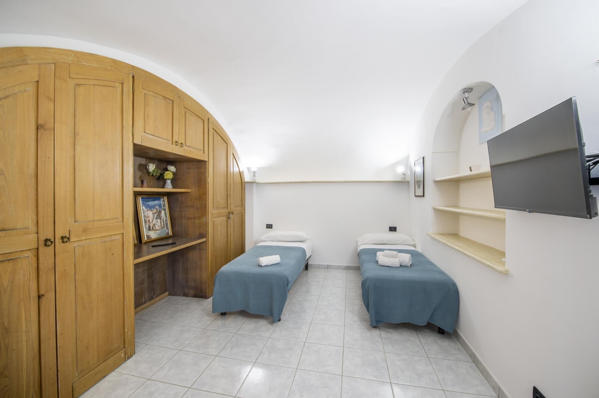A room with two beds, a wooden wardrobe, a TV, and a shelf with decorative items.