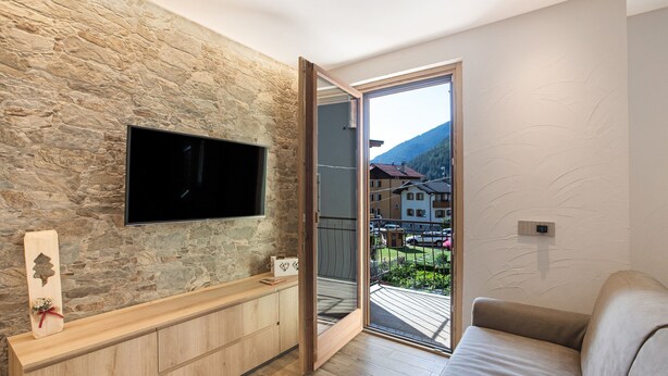 Un salon moderne avec un mur en pierre, un téléviseur à écran plat, une console en bois, un canapé beige et une porte-fenêtre coulissante donnant sur un balcon avec vue sur la montagne.